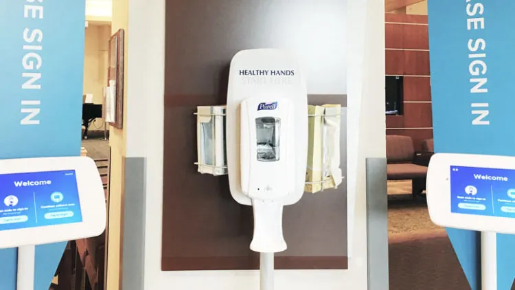 Hand sanitiser dispenser between two Please Sign In check-in kiosks