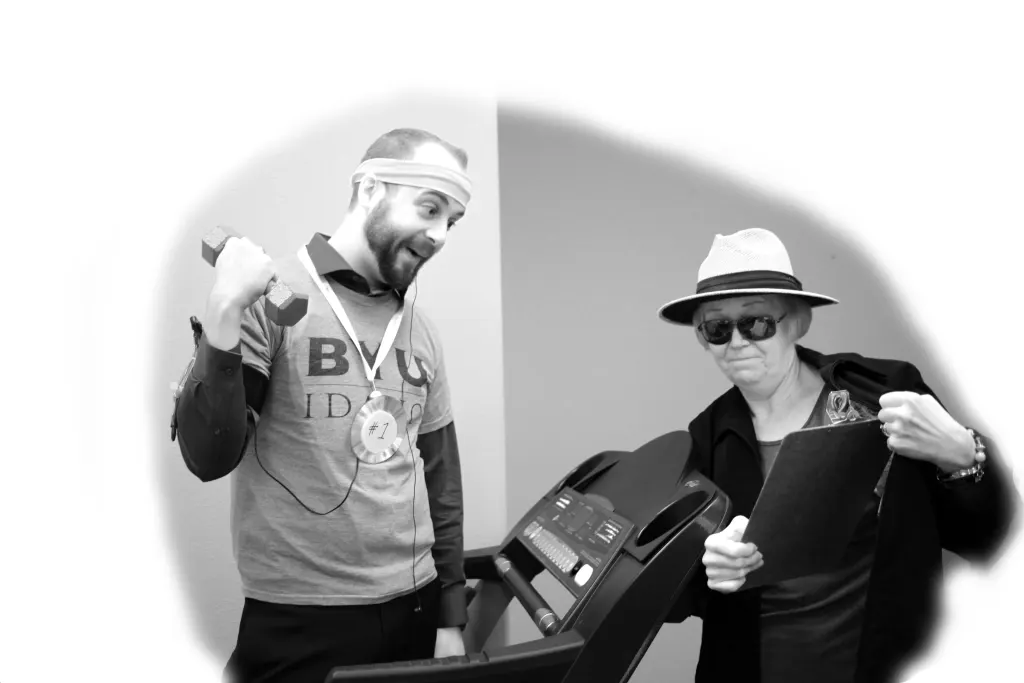 Black-and-white photo of two staff members joking around on a treadmill