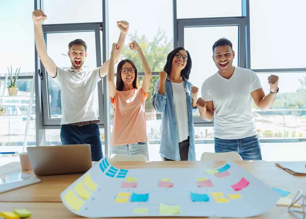 Colleagues cheering with arms raised around a table covered in sticky notes
