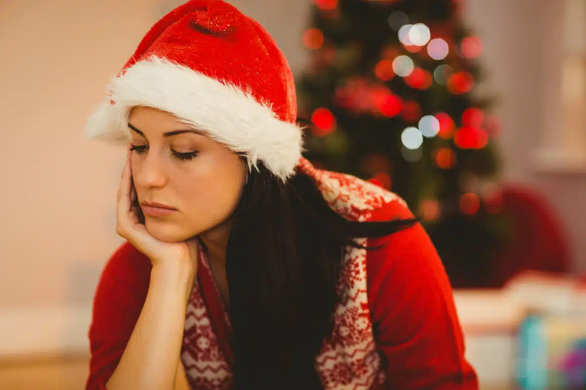 Woman in a Santa hat looking downcast beside a Christmas tree