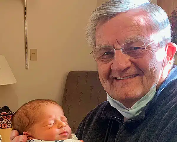 Older man with a mask around his chin holding a sleeping newborn