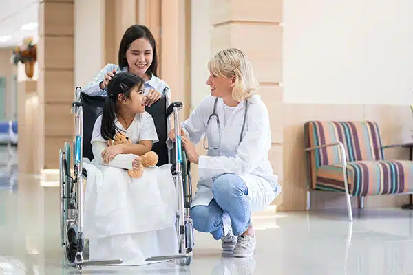 Nurse crouching to talk with a girl in a wheelchair beside her mother