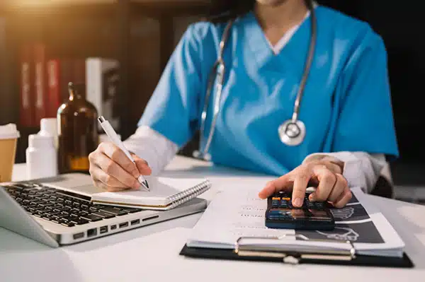 Nurse writing notes at a desk beside a laptop