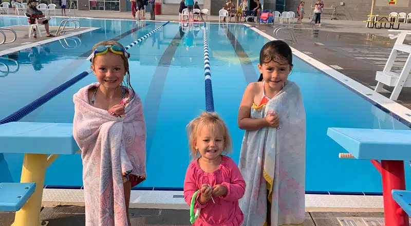 Three young children wrapped in towels beside a swimming pool