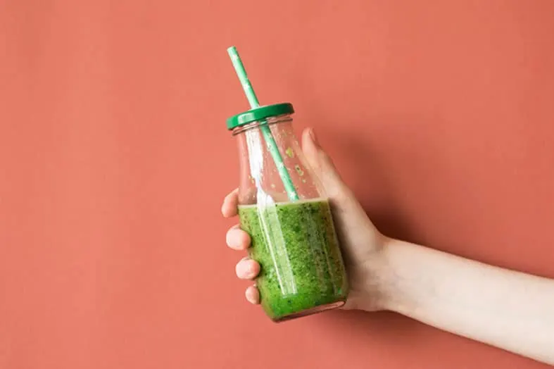 Hand holding a bottle of green smoothie against a coral background