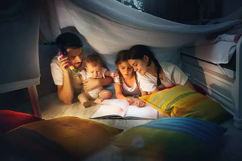Father and children reading by torchlight inside a blanket fort