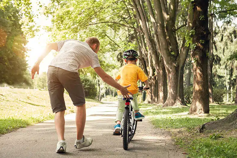 Father helping his son learn to ride a bike along a tree-lined path