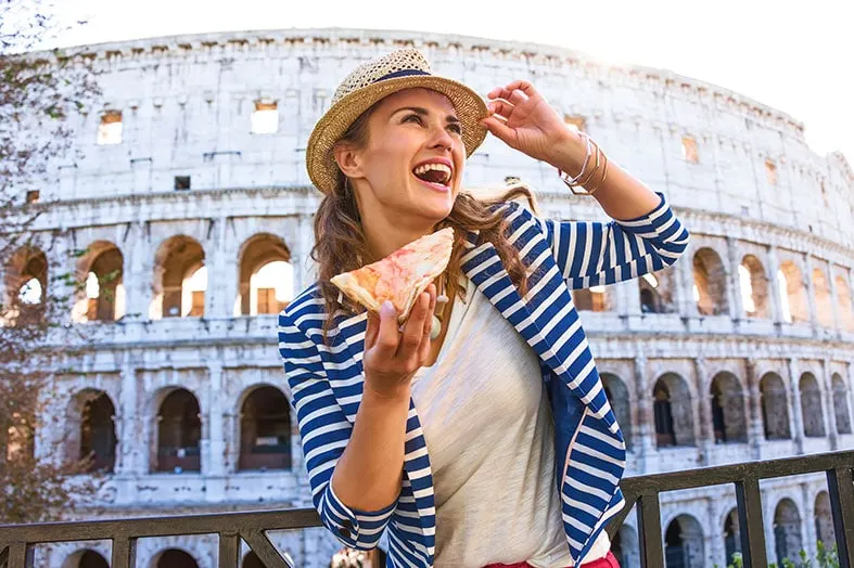 Woman in a sun hat laughing while holding a slice of pizza at the Colosseum