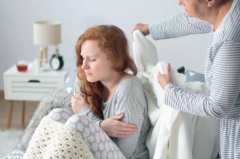 Person resting on a sofa while someone tucks a blanket around them
