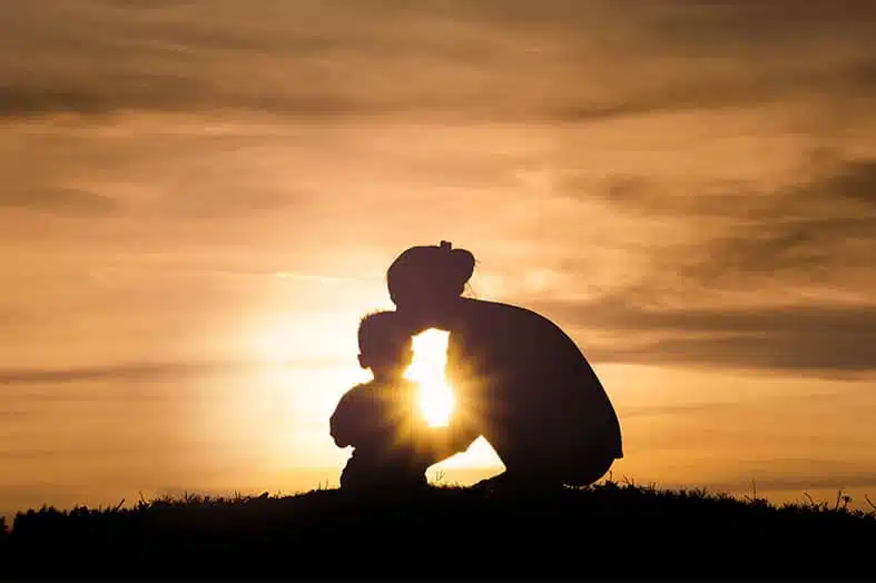 Silhouette of a parent kneeling with a child against a sunset sky
