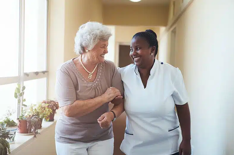 Caregiver walking arm in arm with an older woman down a corridor