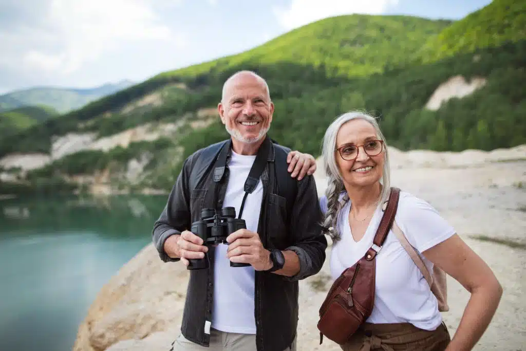 Older couple with binoculars smiling beside a mountain lake