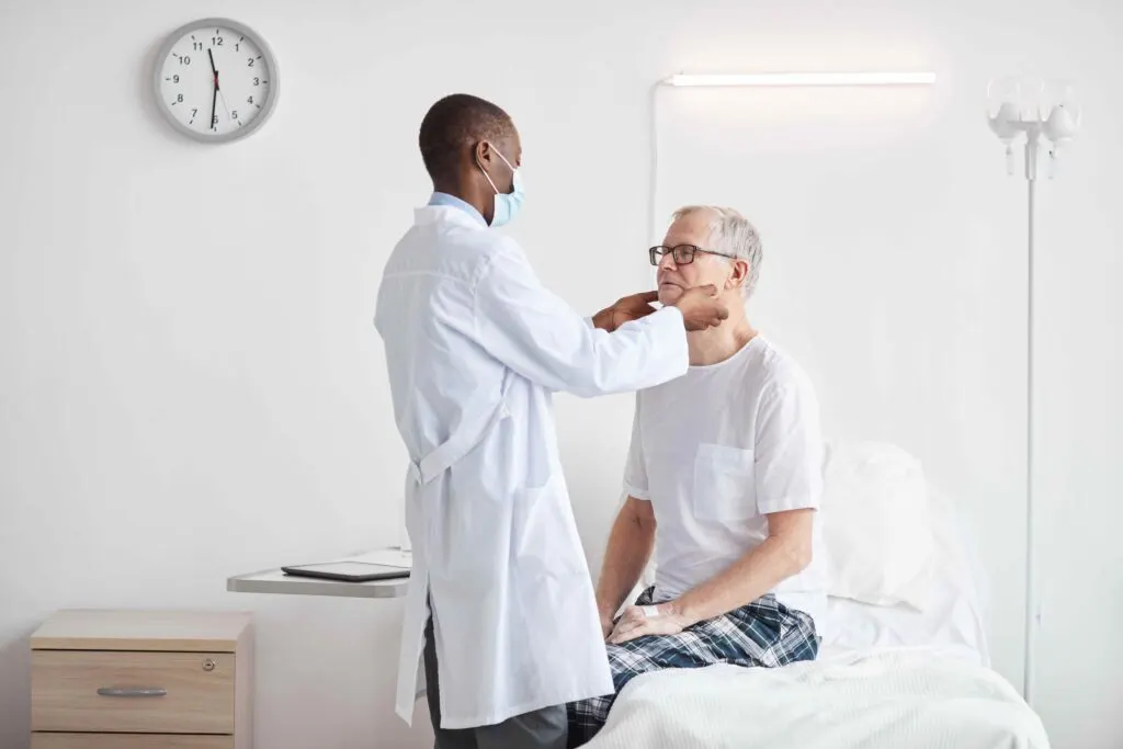 Masked doctor examining an older patient's neck at the bedside