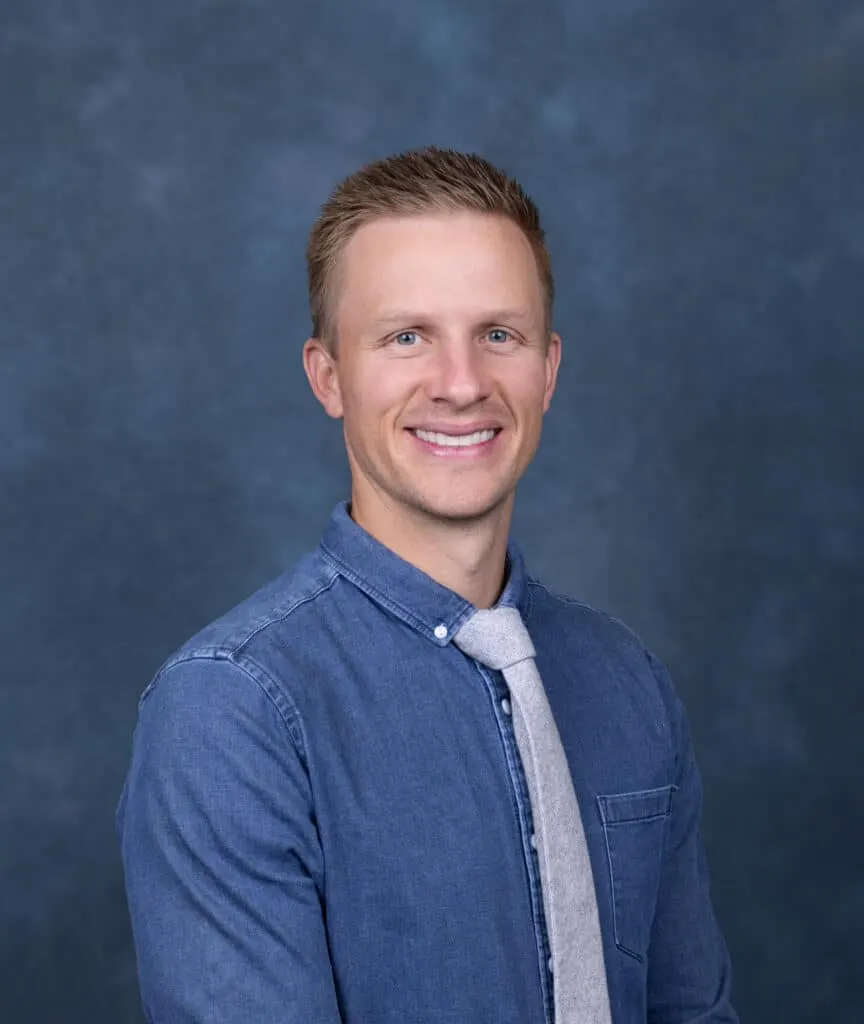 Madisonhealth staff portrait: man in a denim shirt over a grey tee, blue backdrop